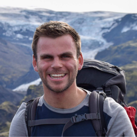 Calum is standing against a mountainous backdrop. He is wearing a large rucksack and is smiling. He has short brown hair.